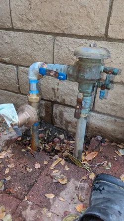Exterior freeze-proof yard hydrant on galvanized riser with a SharkBite PEX push-fit elbow feeding a heavily corroded and rust-stained backflow preventer, mounted at grade against a brick wall with significant oxidation and mineral scaling throughout.