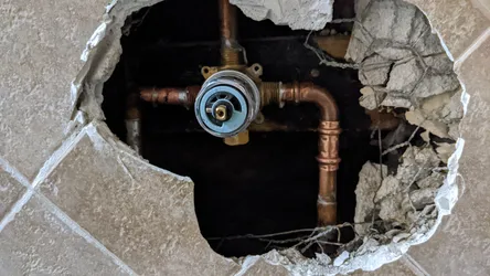 Copper supply lines with sweat-soldered fittings feeding a brass pressure-balancing shower valve, accessed through a demo'd tile wall showing wire lath and mortar bed substrate, no visible corrosion but slight surface oxidation on the copper runs.