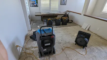 Water damage remediation in progress on a concrete slab subfloor with flooring removed, showing multiple axial air movers and a commercial dehumidifier deployed to dry out a residential living space after a water intrusion event.