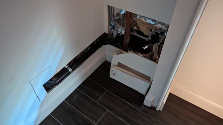 Interior wall opened at baseboard level exposing black ABS or cast iron drain pipe with a banded coupling, visible PVC fittings in the cavity background, batt insulation, foil vapor barrier, and wood framing consistent with a leak investigation or drain re-route in a finished bathroom or hallway.
