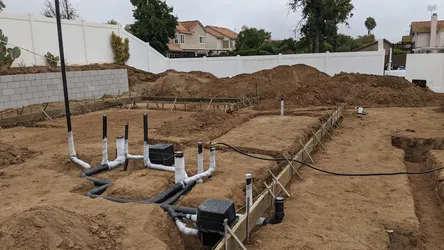 Under-slab rough-in with PVC DWV trunk lines, multiple stub-ups, and two cleanout boxes set in native soil prior to pour, with form boards and rebar stakes visible along the foundation perimeter.