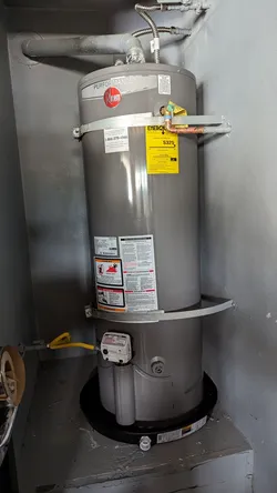 Rheem Performance gas water heater installed in a utility closet on a raised concrete pad, with copper supply connections at the top, yellow CSST flexible gas line at the base, and corrugated stainless flex connectors on the inlet/outlet.