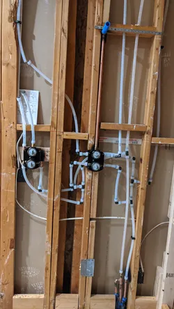 New construction rough-in with white PEX supply lines run through wood-framed studs, two black pressure-balancing shower valves (likely Moen or Delta) with PEX push-fit or crimp connections, and a copper vertical riser stubbed in at right, no visible corrosion or failure modes.