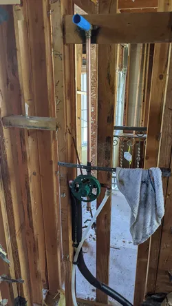 Rough-in shower valve (green-handled, likely Moen Posi-Temp) stubbed out with copper supply pipe capped with a blue PEX end cap, mounted in open stud wall during active remodel with subfloor exposed.