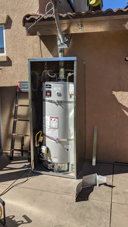 Exterior-installed gas water heater in a dedicated metal enclosure mounted against a stucco wall on a concrete pad, with copper supply connections at the top, a yellow gas flex connector, and a standard T&P relief valve discharge pipe visible.