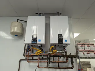 Dual wall-mounted tankless water heaters with copper supply/return manifold, yellow corrugated gas connectors, blue ball valves, and an expansion tank — commercial or utility room install, no visible corrosion but flux residue present on solder joints.