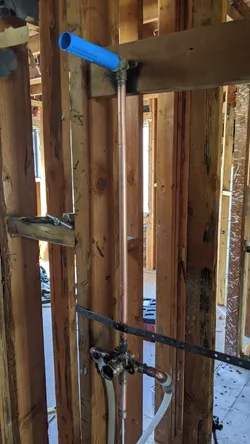 Rough-in interior wall showing a vertical copper riser stubbed to a ball valve at top with a blue CPVC or PEX-AL-PEX horizontal branch, transitioning at the base to what appears to be a shower valve rough-in with copper sweat fittings, framing exposed with no visible corrosion or scale.