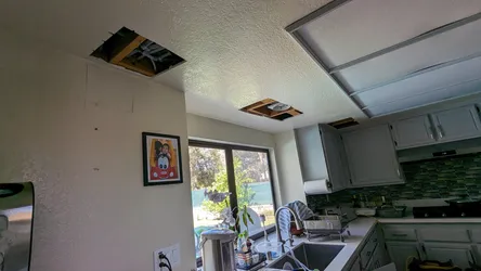 Two ceiling drywall access panels cut open above a kitchen sink area, exposing framing and likely overhead supply or drain lines, consistent with leak investigation or re-pipe access in a single-story residential kitchen.