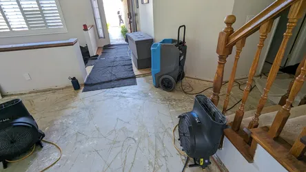 Active water damage mitigation in a residential entryway with flooring removed down to concrete slab, commercial dehumidifiers and air movers deployed, indicating a significant supply or drain line failure beneath the finish floor.