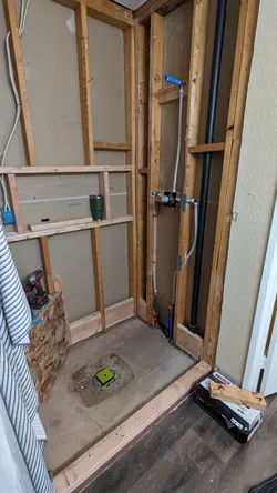 Bathroom rough-in with exposed 2x4 framing showing a PVC vertical drain stack, PEX supply lines with a shower valve rough-in valve body, and a slab floor stub-out for a toilet flange install.