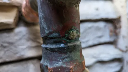Failed copper sweat joint on an exterior or exposed masonry-adjacent line showing active pinhole seepage, heavy verdigris (green oxidation) buildup, and a blown solder bead — classic flux-acid corrosion or improper heat during original solder, needs cut-out and re-sweat or replacement coupling.