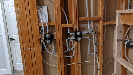 Rough-in wall framing with white PEX tubing routed through studs terminating at two black recessed valve manifold boxes, likely shower or tub rough-in valves, with copper supply visible upper center — no visible corrosion or scale at this stage.