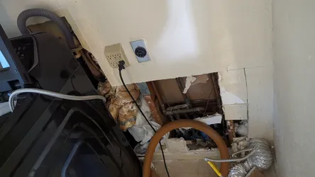 Laundry room wall opened up for water damage remediation, exposing copper supply lines and what appears to be blue PEX in the cavity, with wet/saturated insulation, mold-blackened framing, and a wet-vac hose staged for extraction.