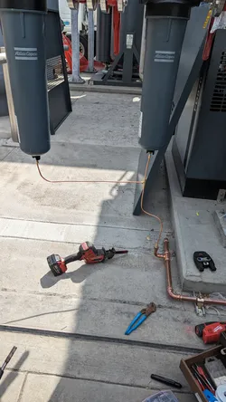 Copper hard-pipe rough-in at slab level serving Atlas Copco commercial air compressors, with press-fit fittings being installed using a Milwaukee press tool; no visible corrosion or scale on new material.