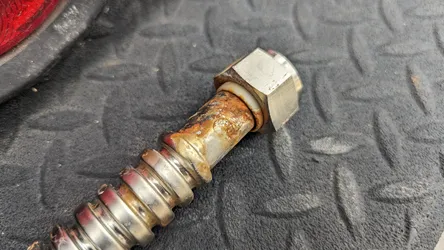 Heavily corroded threaded steel fitting with hex nut transitioning to a braided stainless flexible supply line, showing significant surface rust and galvanic corrosion at the connection interface, removed from service.