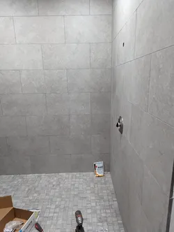 Tile-finished shower enclosure mid-trim-out with large-format gray porcelain wall tile, mosaic floor tile, a brushed-nickel shower valve trim plate installed on the right wall, an overhead rough-in port still open, Moen packaging on site, grout bag visible, no exposed piping or visible failure modes.