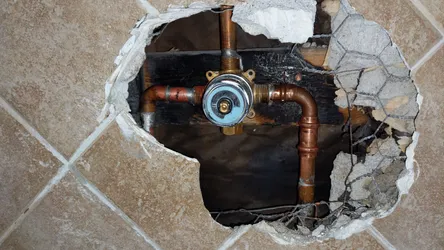 Tile wall opened up to expose copper supply lines with heavy oxidation and mineral staining feeding a brass pressure-balancing shower valve, wire mesh visible in the substrate behind the framing.