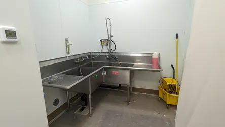 Commercial three-compartment stainless steel sink with wall-mounted pre-rinse spray assembly, chrome supply lines, black ABS drain plumbing below, and an inline water filter on the left wall, in a finished commercial kitchen utility room.