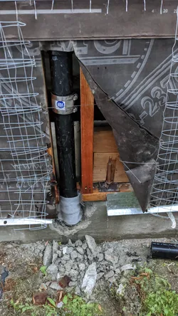Black ABS or cast iron soil stack exposed through a demolished exterior stucco/lath wall at slab level during rough-in access work, with concrete rubble at the base indicating slab saw-cut or demo around the pipe penetration.