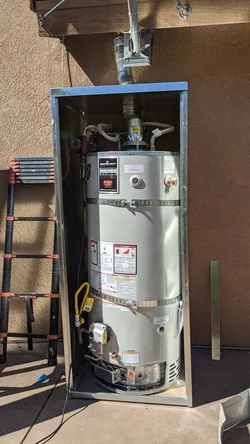 Bradford White gas water heater installed in an exterior stucco-wall alcove with sheet metal enclosure, copper supply connections at top, yellow flexible gas connector at bottom, and B-vent flue exiting through the roof deck above.