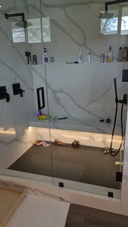 Frameless glass walk-in shower enclosure with matte black trim, dual rain head and handheld fixture setup on large-format porcelain slab surround with integrated bench, linear drain, and recessed niche shelf.
