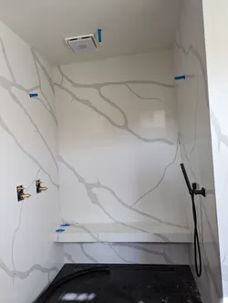 Shower rough-in with brass valve body stubbed out on left wall, blue PEX supply stubs capped at multiple walls and ceiling, matte black handheld holder and linear drain installed, large-format porcelain tile surround mid-installation with open grout joints and bench framed in.