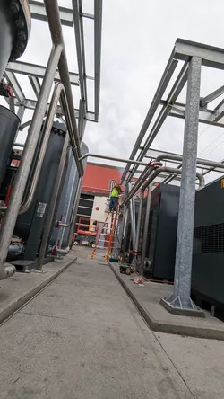 Outdoor commercial mechanical yard with stainless or galvanized steel pipe runs overhead on structural steel unistrut framing, flanking large black industrial tanks or chillers, with a tradesman on a ladder mid-installation on a concrete pad.