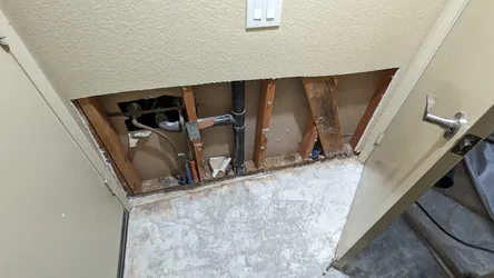 Drywall cut open at ceiling level in what appears to be a garage or utility area, exposing a black ABS drain pipe, copper or galvanized supply lines, and blue PEX stubs running through stud bays, with visible moisture staining on the framing suggesting prior leak damage.