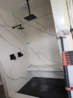 Finish-trim stage walk-in shower with matte black dual rain heads (ceiling-mount and wall-mount), square valve trim, large-format porcelain slab surround, linear drain at the curb, and what appears to be PEX supply still stubbed behind the wall based on the blue fittings visible at mid-wall.