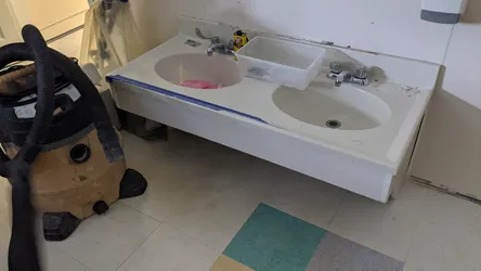 Double-basin cultured marble or cast polymer vanity top with chrome two-handle faucets mid-renovation, supply lines disconnected, blue tape masking the apron, Goof Off solvent on deck, wet-vac staged nearby indicating demo or reseal work in progress.