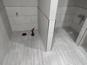 Newly tiled commercial restroom rough-in with copper stub-out on back wall, chrome flush-valve valve body roughed in on right, two floor drains awaiting trim rings, and toilet flange with temporary red wax-ring protector cap still in place.