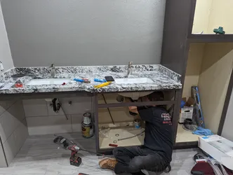 Plumber setting trim on a new double-vanity install in a finished bathroom — braided stainless supply lines visible, brushed nickel faucets already set in granite top, drain assembly being fitted with PVC P-trap, no corrosion or prior failures present.