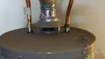 Gas water heater top connections showing corrugated copper flexible supply lines on hot and cold ports with brass fittings, a sheet metal draft hood with foil-taped flue collar junction showing improper tape repair, and heavy dust accumulation on the tank top.