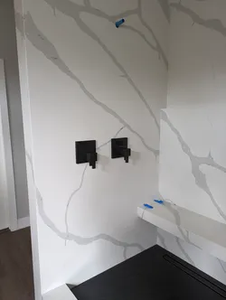 New construction shower trim-out with two matte black volume/diverter valves set into large-format porcelain or quartz slabs, blue PEX stub-outs capped at the bench and overhead rain-head location, linear drain at the curb — finish stage, no visible corrosion or failure modes.