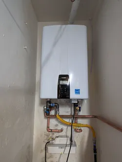 Navien tankless water heater wall-mounted in a utility alcove, plumbed with copper supply lines and a corrugated stainless gas flex connector, PVC condensate drain running overhead, with isolation valves and a pressure relief assembly at the unit connections.