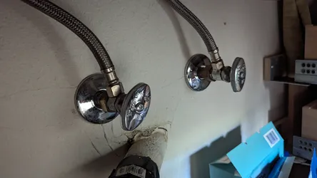 Two chrome angle stop shut-off valves with braided stainless supply lines stubbing out of a textured drywall finish, showing minor water staining and efflorescence at the lower escutcheon, consistent with a bathroom sink or laundry rough-in.
