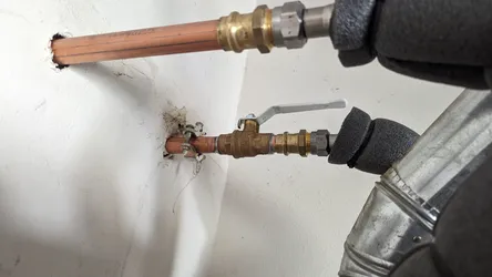 Copper supply line stubbed through drywall with a brass ball valve and compression/push-fit fittings on both ends, foam-insulated flex connector on the outlet side, pipe clamp at the wall penetration, no visible corrosion.