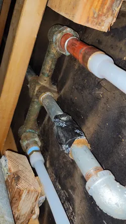 Corroded copper supply lines with heavy verdigris patina transitioning to PEX repipe in a crawlspace or subfloor rough-in, with a temporary rubber repair clamp (Fernco or similar) bandaging an active leak point on the copper main.