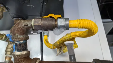 Gas appliance connection at a furnace or water heater showing a corroded black iron pipe elbow with a brass ball valve transitioning to a yellow CSST flexible gas connector, with active soap bubble leak test visible at the union fitting.