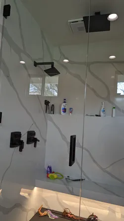 Finished walk-in shower with matte black trim kit — wall-mount rain head, dual volume/diverter valves, and linear drain — set into large-format porcelain slab surround with frameless glass enclosure; no visible supply rough-in or failure modes.