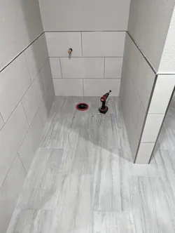 Newly tiled bathroom rough-in with a copper stub-out protruding from the wall tile and a black ABS closet flange set in the floor tile, grout lines still open, finish fixtures not yet installed.
