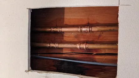 Two parallel copper supply lines with soldered sweat couplings showing oxidation and surface tarnish, accessed through a drywall cutout revealing wood framing, with a gray electrical conduit or PVC line running below between floor joists.