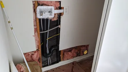 Interior wall opened up exposing a black ABS drain stack with a P-trap elbow, gray PEX supply lines, flexible metallic conduit, and a new outlet box mounted to studs, with fiberglass batt insulation visible and water damage staining on the tile floor indicating a prior leak event.