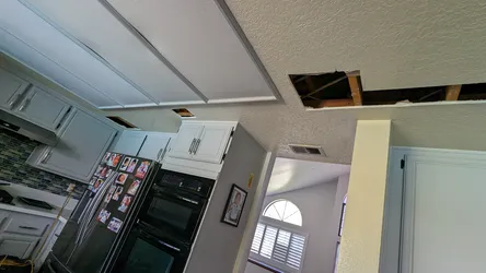 Residential drywall ceiling cut open for leak investigation above a kitchen/living area, with framing and subfloor visible in the access hole, indicating active water intrusion from above requiring trace and repair of supply or drain line running through the floor/ceiling assembly.