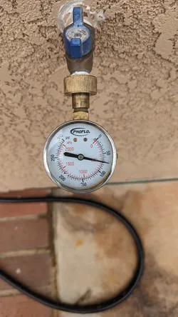 ProFlo brass hose-thread pressure gauge screwed onto an exterior hose bib with a blue quarter-turn ball valve, reading approximately 75 psi static supply pressure against a stucco exterior wall.