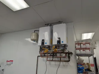 Dual tankless water heaters wall-mounted in a commercial utility room, plumbed in copper with yellow CSST gas flex connectors, a single expansion tank shared between units, and no visible corrosion or scale on the copper manifold assembly.