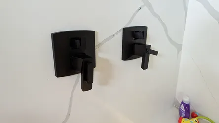 Two matte black in-wall valve trim kits — left appears to be a pressure-balance shower valve with diverter, right a volume/diverter control — mounted on tile surround in a finished bathroom, no visible corrosion or supply connections exposed.