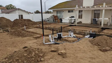 Slab rough-in for a residential addition showing ABS or PVC DWV stubouts with cleanouts and black vertical riser pipes laid out in trenched soil prior to concrete pour, no visible corrosion or pressure supply lines yet.
