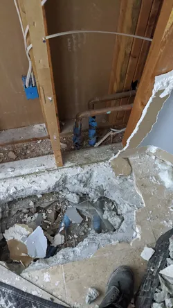 Slab demo in progress with tile and concrete broken out to expose underslab rough-in, PEX supply lines with blue crimp fittings visible through the stud wall, concrete debris cleared for drain access or reroute.