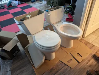 Two vitreous china two-piece toilets staged on cardboard in a living area — tanks removed, seats still on — prepped for installation, no supply lines or wax rings visible yet.